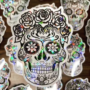 May include: A holographic sticker featuring a black and white sugar skull with floral details. The skull has a diamond on its forehead and a heart on its chin. The background is a rainbow iridescent color.