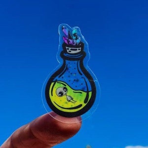May include: A clear sticker with a green, yellow, and blue potion bottle with a small alien inside. The bottle has a black lid and iridescent crystals on top.