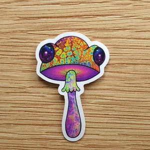Puede incluir: Una colorida pegatina de hongo con un dise&ntilde;o psicod&eacute;lico. El sombrero del hongo presenta patrones en naranja, amarillo y verde, con acentos morados y dos grandes ojos morados con forma de galaxia. El tallo es morado y la pegatina est&aacute; sobre una superficie de madera.