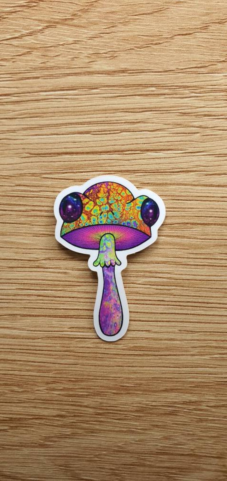 Puede incluir: Una colorida pegatina de hongo con un dise&ntilde;o psicod&eacute;lico. El sombrero del hongo presenta patrones en naranja, amarillo y verde, con acentos morados y dos grandes ojos morados con forma de galaxia. El tallo es morado y la pegatina est&aacute; sobre una superficie de madera.