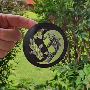 May include: A round black sticker featuring a Yin and Yang design with two koi fish. One fish is light green, the other is black and white. The sticker is held in front of a green leafy background.
