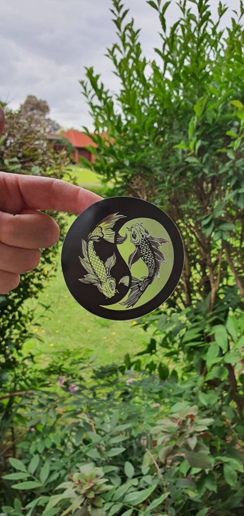 May include: A round black sticker featuring a Yin and Yang design with two koi fish. One fish is light green, the other is black and white. The sticker is held in front of a green leafy background.