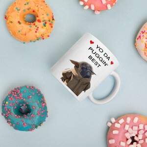May include: White ceramic mug with a brown and black pug dressed as Yoda. The mug has red hearts and the text "Yo Da Puggin' Best" printed on it.
