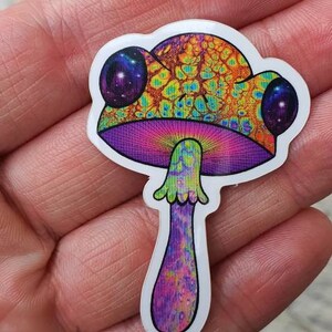 Puede incluir: Una colorida pegatina con un dise&ntilde;o de hongo psicod&eacute;lico. El hongo tiene un sombrero naranja y amarillo vibrante con un patr&oacute;n texturizado, branquias moradas y un tallo morado y verde. La pegatina tiene un borde blanco.
