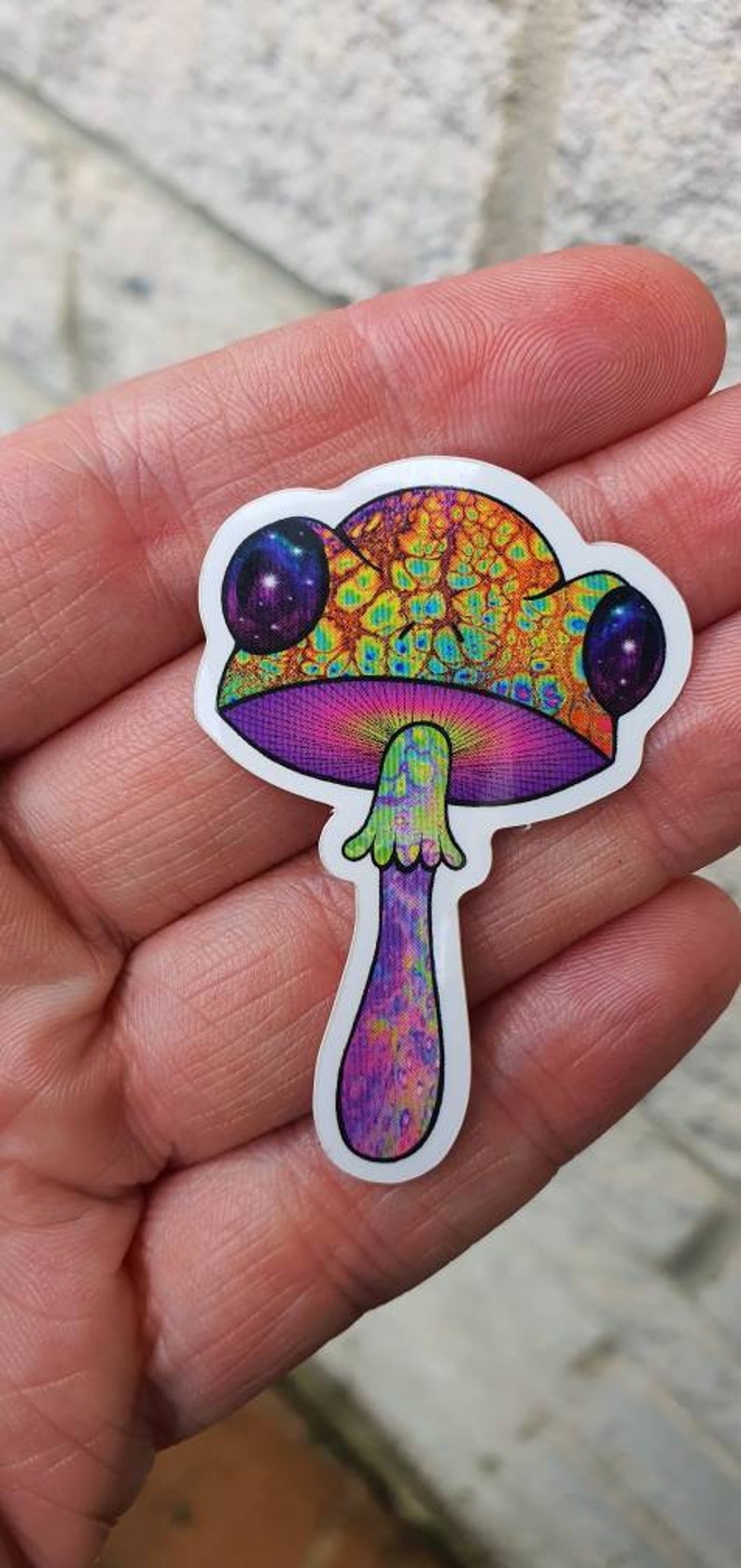 Puede incluir: Una colorida pegatina con un dise&ntilde;o de hongo psicod&eacute;lico. El hongo tiene un sombrero naranja y amarillo vibrante con un patr&oacute;n texturizado, branquias moradas y un tallo morado y verde. La pegatina tiene un borde blanco.
