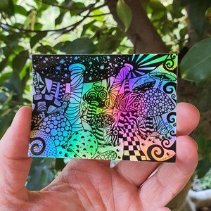 May include: A holographic sticker with a psychedelic design featuring colourful mushrooms and swirls.