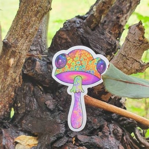 Puede incluir: Una colorida pegatina de hongo con un dise&ntilde;o psicod&eacute;lico. La pegatina presenta un tallo morado, un sombrero rosa y naranja y grandes ojos morados. La pegatina est&aacute; colocada en una rama de &aacute;rbol.