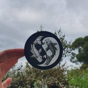 May include: A black and white Yin and Yang koi fish design on a round sticker. The sticker features two koi fish in a circular pattern, with one fish white and the other black. The background is a cloudy sky and green foliage.