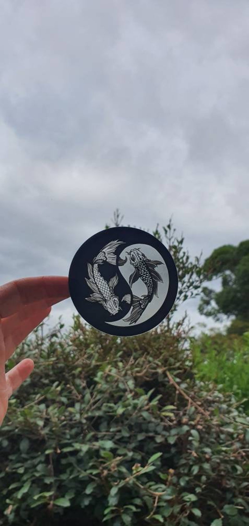 May include: A black and white Yin and Yang koi fish design on a round sticker. The sticker features two koi fish in a circular pattern, with one fish white and the other black. The background is a cloudy sky and green foliage.