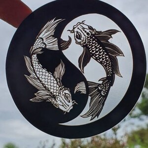 May include: A black and white Yin and Yang design featuring two koi fish. The fish are intricately detailed with scales and fins, set against a cloudy sky background. The artwork is circular.