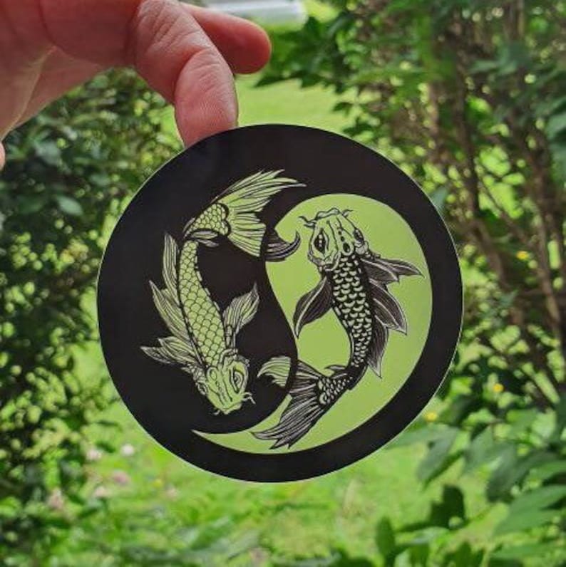May include: Round black sticker featuring a Yin and Yang design with two koi fish. The fish are light green with black details, set against a black background with a light green crescent. The sticker is held up by a hand.