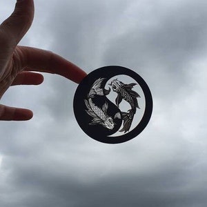 May include: A black and white Yin and Yang design featuring two koi fish. The circular design is held up against a cloudy sky. The fish are detailed with scales and fins, creating a balanced and symbolic image.