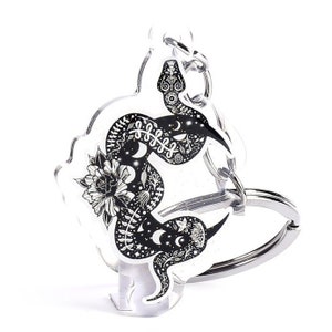 May include: A black and white snake with intricate details, including moons and flowers, is printed on a clear acrylic keychain. The keychain has a silver metal ring.