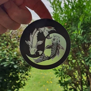 May include: A round black sticker featuring two koi fish in a yin-yang design. The fish are white with detailed scales and fins, set against a light green background. The sticker is held up against a backdrop of green foliage.