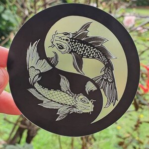 May include: A round black sticker featuring a Yin and Yang design with two koi fish. The fish are detailed with scales and fins, set against a light green background.
