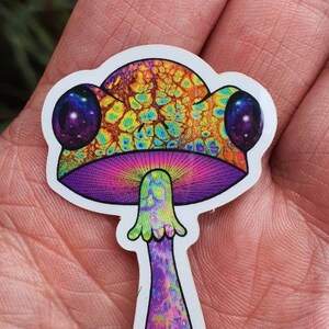 Puede incluir: Una colorida pegatina de hongo con un dise&ntilde;o psicod&eacute;lico. El sombrero del hongo es una mezcla de amarillo, naranja y verde, con dos grandes ojos morados oscuros. El tallo es morado con un patr&oacute;n texturizado.