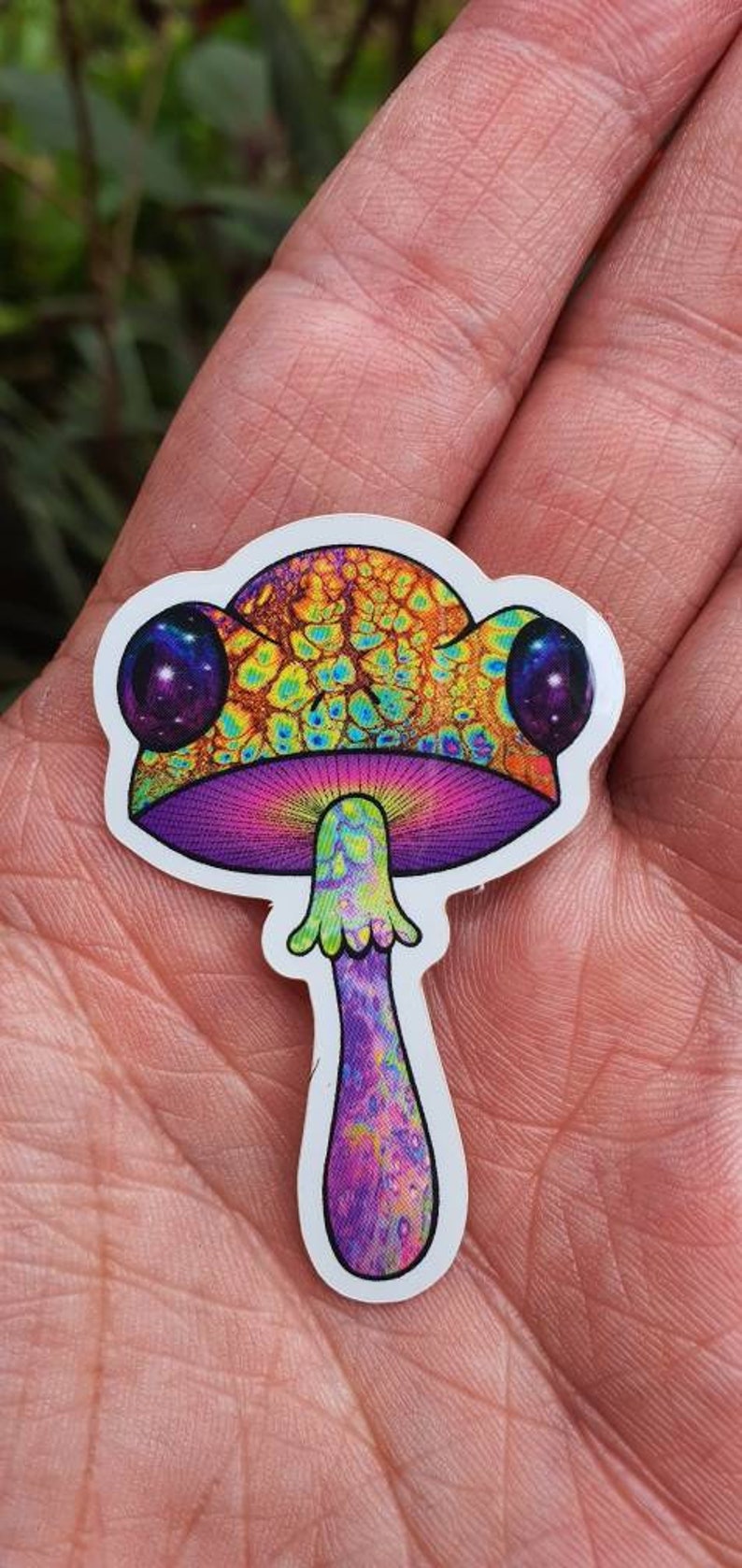 Puede incluir: Una colorida pegatina de hongo con un dise&ntilde;o psicod&eacute;lico. El sombrero del hongo es una mezcla de amarillo, naranja y verde, con dos grandes ojos morados oscuros. El tallo es morado con un patr&oacute;n texturizado.