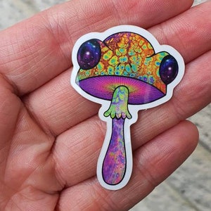 Puede incluir: Una colorida pegatina de hongo con un dise&ntilde;o psicod&eacute;lico. El sombrero del hongo presenta un patr&oacute;n texturizado en tonos naranja, amarillo y verde. El tallo es morado con un patr&oacute;n texturizado. Dos grandes ojos redondos con tem&aacute;tica de galaxia est&aacute;n en el sombrero.