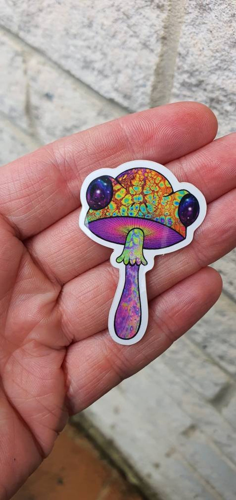 Puede incluir: Una colorida pegatina de hongo con un dise&ntilde;o psicod&eacute;lico. El sombrero del hongo presenta un patr&oacute;n texturizado en tonos naranja, amarillo y verde. El tallo es morado con un patr&oacute;n texturizado. Dos grandes ojos redondos con tem&aacute;tica de galaxia est&aacute;n en el sombrero.