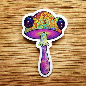 Puede incluir: Una colorida pegatina con un dise&ntilde;o de hongo psicod&eacute;lico. El sombrero del hongo tiene un patr&oacute;n texturizado en tonos de amarillo, azul y naranja. El tallo es morado y verde, con un dise&ntilde;o de galaxia en los ojos.