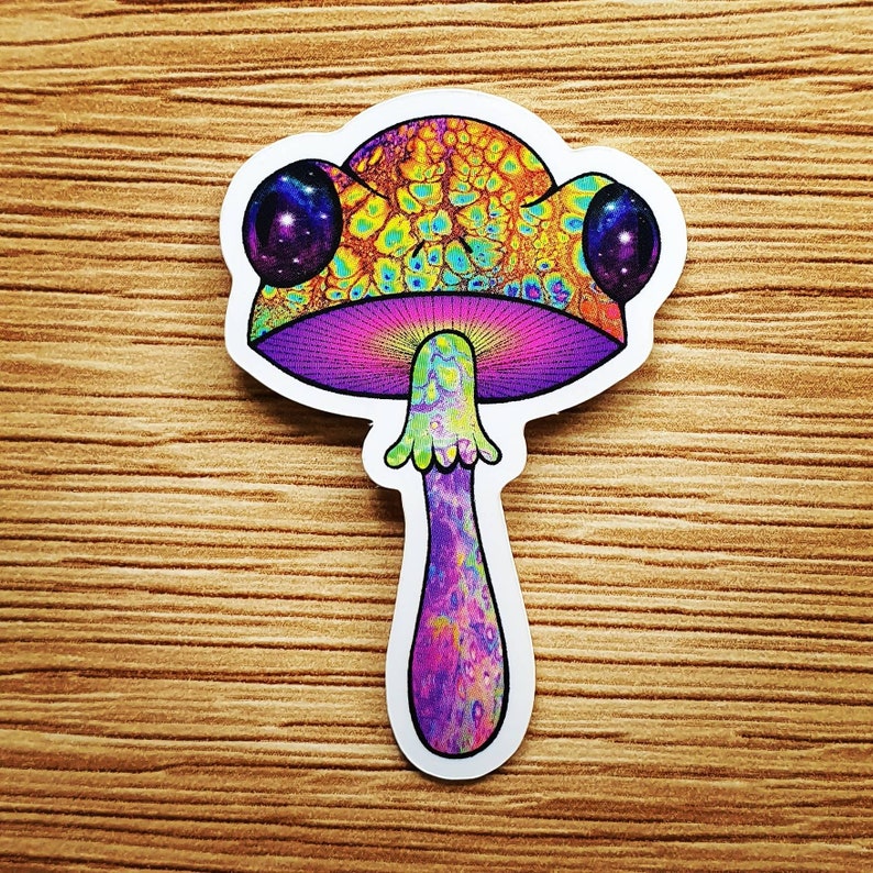 Puede incluir: Una colorida pegatina con un dise&ntilde;o de hongo psicod&eacute;lico. El sombrero del hongo tiene un patr&oacute;n texturizado en tonos de amarillo, azul y naranja. El tallo es morado y verde, con un dise&ntilde;o de galaxia en los ojos.