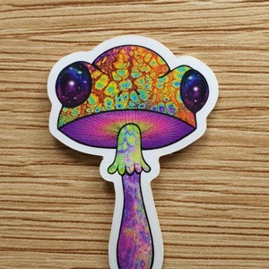 Puede incluir: Una colorida pegatina de una seta psicod&eacute;lica. El sombrero de la seta presenta una vibrante mezcla de naranja, amarillo y verde, con dos c&iacute;rculos p&uacute;rpuras en forma de galaxia. El tallo es morado y rosa.