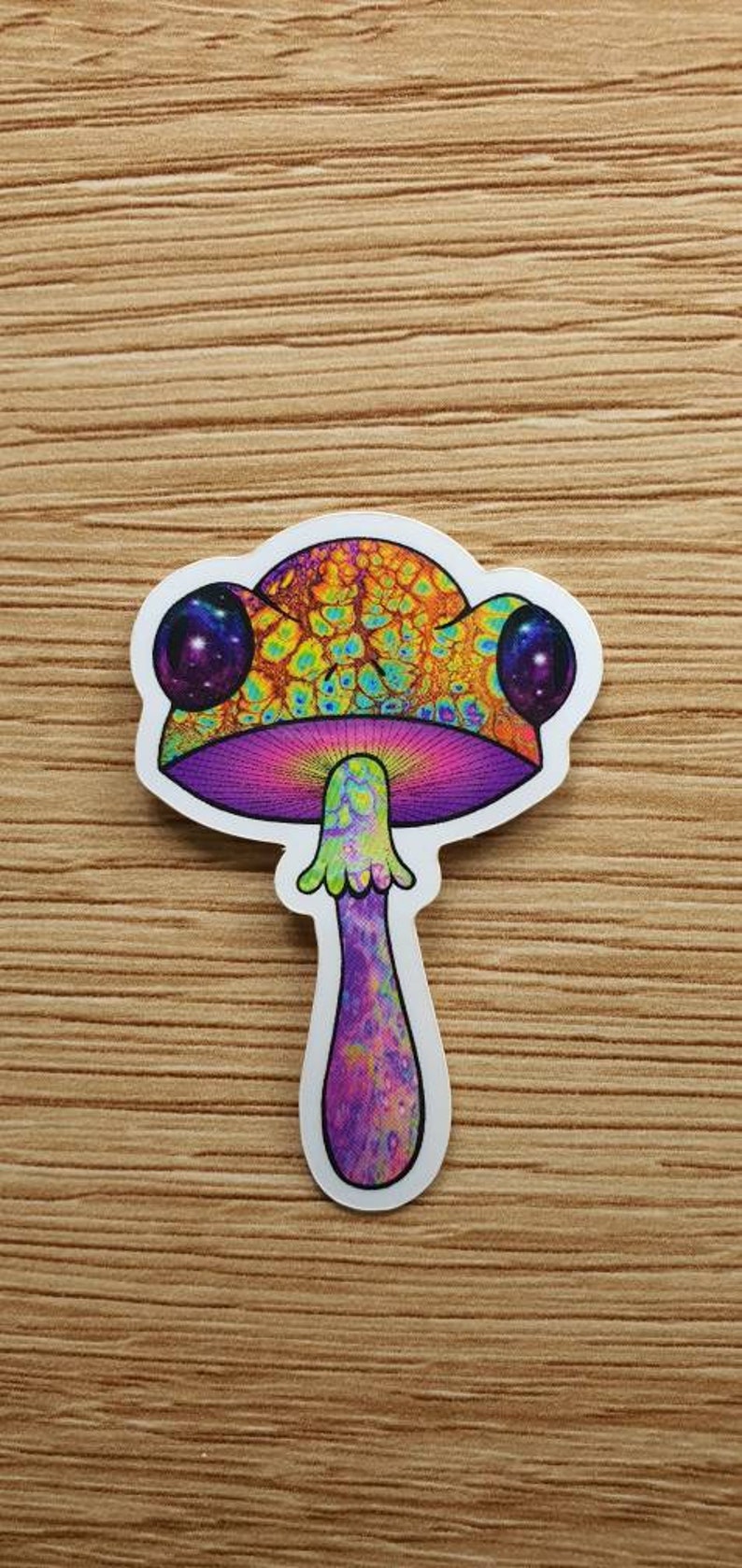 Puede incluir: Una colorida pegatina de una seta psicod&eacute;lica. El sombrero de la seta presenta una vibrante mezcla de naranja, amarillo y verde, con dos c&iacute;rculos p&uacute;rpuras en forma de galaxia. El tallo es morado y rosa.