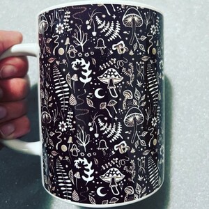 May include: A white ceramic mug with a black and white design of mushrooms, ferns, and other forest elements. The design is intricate and detailed, and the mug is perfect for a hot beverage.
