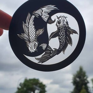 May include: A black and white circular sticker featuring two koi fish in a yin and yang design. The fish are detailed with scales and fins, set against a white background within a black circle. The sticker is held up against a cloudy sky.
