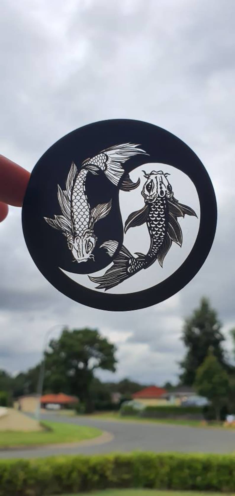 May include: A black and white circular sticker featuring two koi fish in a yin and yang design. The fish are detailed with scales and fins, set against a white background within a black circle. The sticker is held up against a cloudy sky.