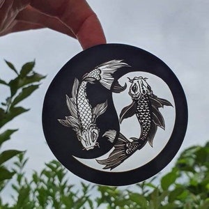 May include: A black circular sticker featuring a white yin and yang design with two koi fish. The fish are detailed with scales and fins, creating a balanced and symmetrical composition. The sticker is held against a backdrop of green foliage and a cloudy sky.
