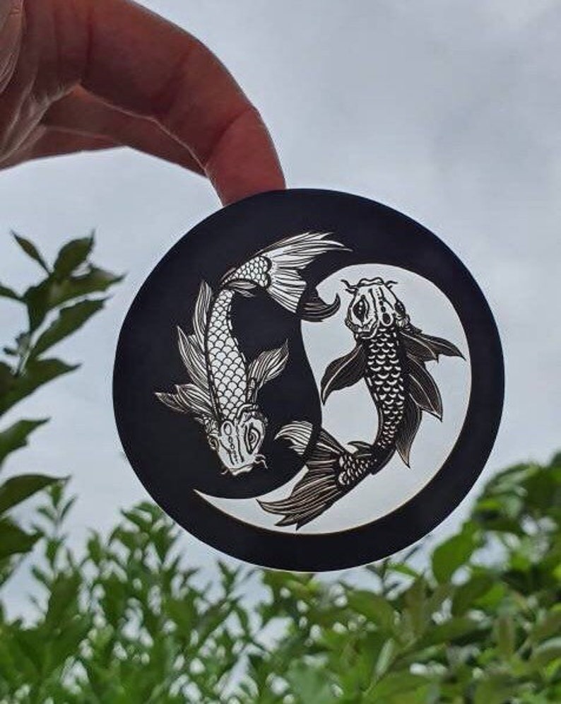 May include: A black circular sticker featuring a white yin and yang design with two koi fish. The fish are detailed with scales and fins, creating a balanced and symmetrical composition. The sticker is held against a backdrop of green foliage and a cloudy sky.