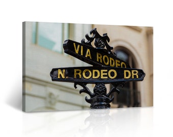 May include: A black metal street sign with gold lettering pointing in different directions. The sign reads "Via Rodeo" and "N. Rodeo Dr."