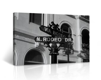 May include: Black and white photo of a street sign with the words "Via Rodeo" and "N. Rodeo Dr." on it. The sign is mounted on a black pole and is in front of a building with arched windows.