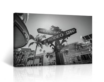 May include: Black and white photo of a street sign with the words "Via Rodeo Dr" and "N. Rodeo Dr" on it. The sign is in front of a building with palm trees in the background.