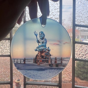 May include: A round glass ornament featuring a blue statue of Neptune holding a trident, set against a beach scene with a sunset sky. The ornament is held by a hand with a blue ribbon for hanging.