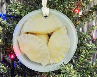 May include: A white ceramic ornament shaped like a bowl filled with four white dumplings. The ornament is hanging from a white ribbon on a green Christmas tree.