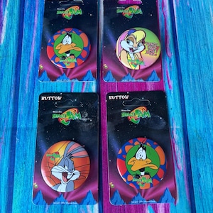 May include: Four Space Jam badges on black cards. Each badge features a different Looney Tunes character: Daffy Duck, Lola Bunny, Bugs Bunny, and Daffy Duck. The badges have colourful backgrounds and the Space Jam logo. The cards are on a blue wooden surface.