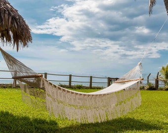 May include: A white, woven hammock with wooden support bars hangs on a grassy lawn. The hammock has a fringed edge and is suspended between two points. The background includes a fence, ocean, and a cloudy sky.
