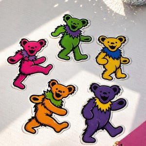May include: Five colorful dancing bear stickers. Each bear is a different color: pink, green, yellow, orange, and purple. They all have a smiling face and a ruffled collar. The stickers are on a white surface.