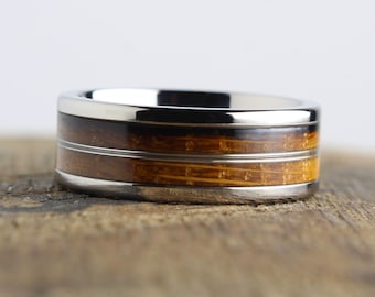 Puede incluir: Un anillo de titanio plateado con dos bandas de incrustaciones de madera. La madera es de color marrón claro con una veta marrón más oscura.