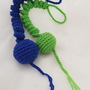 Puede incluir: Un juguete de crochet de color azul y verde. El juguete está hecho de dos bolas conectadas por un cordón largo. Las bolas están hechas de hilo de crochet tejido apretado.