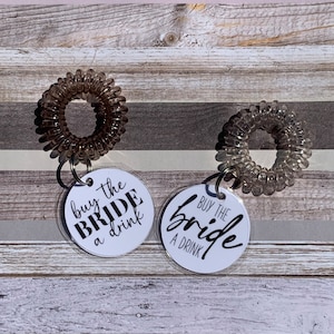 May include: Two clear spiral hair ties with silver key rings attached to white circular tags. The tags have the phrase "buy the bride a drink" in black script. The background is a mix of wood and gray stripes.