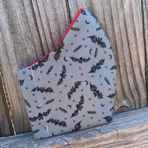 Puede incluir: Una mascarilla facial de tela gris con un patrón repetido de murciélagos negros y estrellas blancas. La mascarilla tiene un borde rojo.