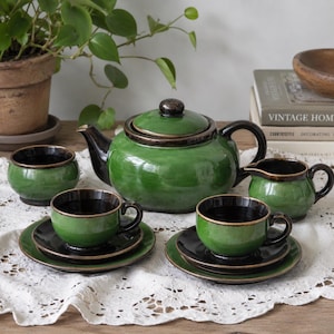 May include: A green ceramic tea set with black detailing. The set comprises a teapot, creamer, sugar bowl, and two teacups with saucers. The teapot has a dark brown handle and lid. The teacups and saucers are black with green rims.