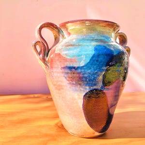 May include: A ceramic vase with two ornate, curled handles and a wide rim. The surface features a glossy, multi-coloured glaze with blended patches of vibrant blue, deep green, warm orange, and soft pink, creating a fluid, abstract appearance.