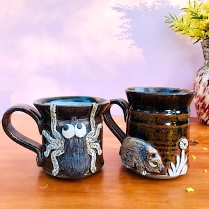 May include: Two dark brown ceramic mugs with glossy finishes. The left mug features a raised, textured spider with large white eyes. The right mug displays a sculpted brown creature resembling a rodent and white floral accents near the base.