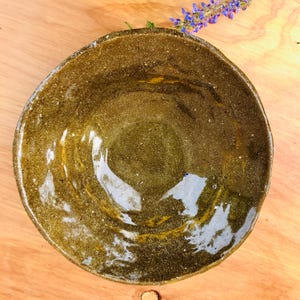 May include: A round, handmade ceramic bowl with an uneven, organic rim. The interior features a glossy, dark olive-green glaze with subtle golden-brown speckles and swirling patterns, reflecting light across its textured, slightly undulating surface.