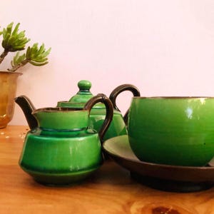 May include: A ceramic tea set featuring a vibrant green glaze with dark brown trim. The set includes a small creamer, a teapot with a lid, and a rounded teacup resting on a matching dark brown saucer, all showcasing a smooth, glossy finish.