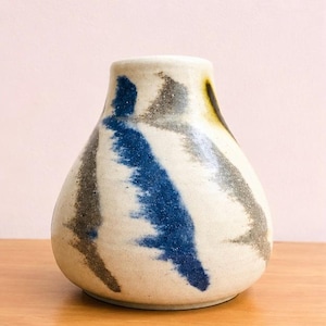 May include: A ceramic vase with a rounded, tapered shape and a wide base. The surface is off-white with a speckled texture, featuring bold, abstract brushstrokes in deep blue and muted grey that wrap around the body of the vessel.